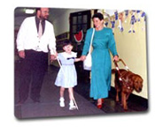 Cane teacher, child with cane, and blind adult with guide dog travel down a school hall together